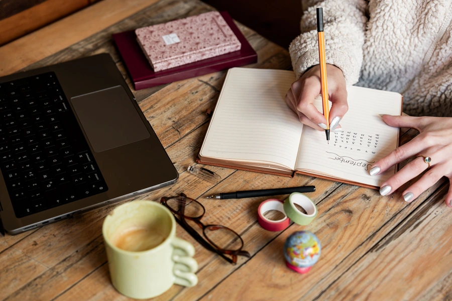Manos de una mujer trabajando con lápiz en un cuaderno junto a un portátil y materiales de escritorio